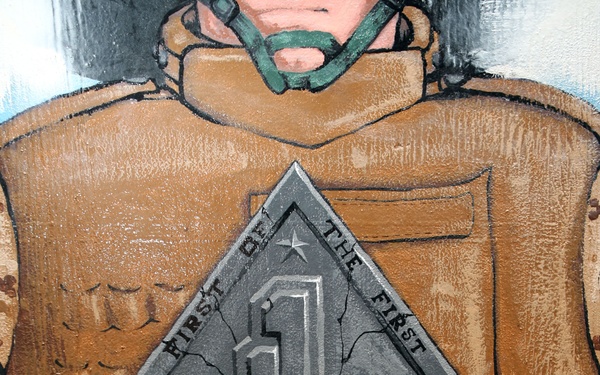 A painted mural or artwork depicting a military figure in combat gear pulling open their uniform to reveal a diamond-shaped insignia bearing the number '1' and the text 'First of the First'. A red ribbon banner below reads 'Ready To Fight'. The style is illustrative and bold, consistent with military unit pride artwork.