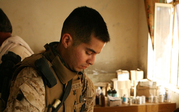 A U.S. military servicemember in MARPAT desert camouflage uniform and tactical vest is examining and cataloging pharmaceutical supplies on a table. The setting appears to be an indoor room, possibly a medical facility or aid station, with numerous medication bottles and boxes spread across multiple tables. The servicemember's name tape reads 'ARREDONDO' and they appear to be conducting a medical supply inventory or assessment.