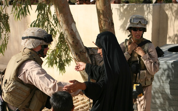 The image shows two military personnel in desert camouflage uniforms and combat gear interacting with a woman in a black abaya and a young child holding a red stuffed animal. The soldier on the left appears to be handing something to or gesturing toward the woman, while a second soldier stands in the background. The setting appears to be an outdoor area with sandy ground, a tree, and a light-colored wall.