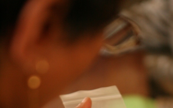 A person wearing military digital camouflage uniform is holding a small clear zip-lock plastic bag with handwritten Arabic script on it. The individual also holds a dark pen or marker in the same hand. The image is partially blurred with the focus centered on the bag and hands.