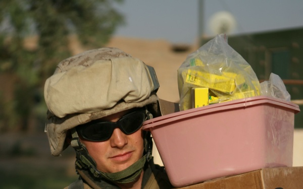 A U.S. Marine in desert MARPAT camouflage uniform, combat helmet, and tactical vest is carrying a stack of boxes and a pink plastic bin filled with supplies. One of the cardboard boxes is branded with 'GSK GlaxoSmithKline' and marked 'RX' in black marker, suggesting pharmaceutical or medical supplies. The setting appears to be an outdoor area in a Middle Eastern environment based on the architecture and vegetation visible in the background.