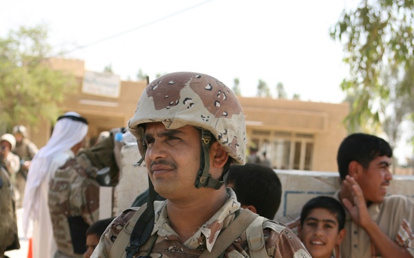 A soldier in desert camouflage uniform and helmet stands in the foreground holding a Garrett handheld metal detector, suggesting a security checkpoint or screening operation. Several civilians, including children and adults in traditional Middle Eastern dress, are visible in the background along with what appears to be another uniformed soldier. The setting appears to be an outdoor area near a tan/beige building, consistent with a Middle Eastern environment.