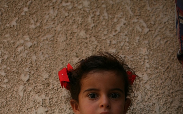 A young girl, approximately 2-4 years old, sits against a rough textured beige wall. She is dressed in a mint green ruffled dress with two red hair bows and appears to be holding a stuffed toy. Her expression is serious and direct, looking straight into the camera.