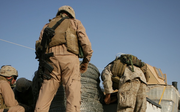 Military personnel in desert camouflage uniforms are working on the flatbed of a military vehicle, handling a large coil of concertina/razor wire. The scene appears to be in an arid environment, likely a forward operating base or staging area. At least three soldiers are visible, engaged in loading or unloading operations.