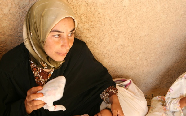 A woman wearing a black abaya and olive-green hijab sits on a floor against a textured wall, holding a sleeping infant and a piece of white bread or flatbread. Two other children are visible nearby, one older girl in a white floral dress sitting to the right holding markers. The setting and clothing suggest a Middle Eastern context, possibly a shelter or temporary dwelling.