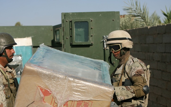 Two soldiers in desert camouflage uniforms are handling a large cardboard box wrapped in plastic sheeting. They appear to be in an outdoor area with a military vehicle, brick wall, and palm trees visible in the background. The setting appears to be a Middle Eastern environment based on the architecture and vegetation.
