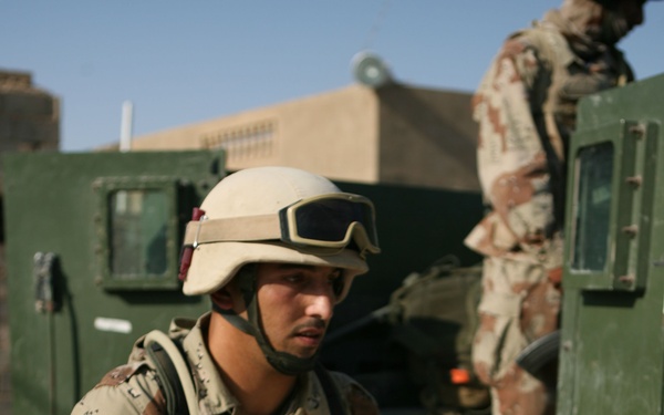 A soldier wearing desert camouflage uniform, tactical vest, and helmet with mounted goggles is handling a large white plastic crate or cooler wrapped in plastic. A second soldier in matching camouflage is visible in the background near what appears to be a military vehicle (HMMWV/Humvee). The setting appears to be an arid, urban environment based on the sandy-colored building and clear blue sky visible in the background.