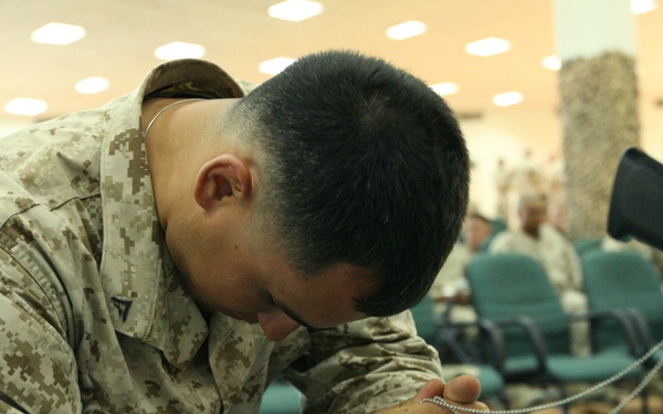 A U.S. Marine in desert MARPAT camouflage uniform, identified by the name tape 'PEREZ', is kneeling in prayer with head bowed and hands clasped together holding dog tags. The setting appears to be a military chapel or religious service space, with other service members visible seated in the background. The scene conveys a moment of personal religious observance during a military chapel service.
