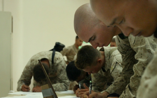 A group of military personnel in desert camouflage uniforms are leaning over a long table, writing or filling out documents. The scene appears to be an administrative or testing session conducted in a sparse indoor facility. Multiple forms with visible grid or chart layouts are spread across the table surface.