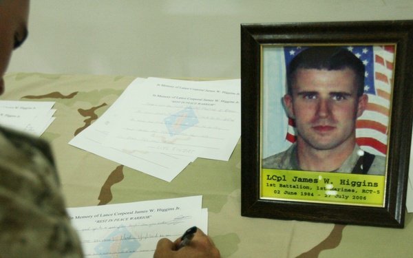 A memorial scene showing a framed photograph of Lance Corporal James W. Higgins, 1st Battalion, 1st Marines, RCT-3, born 02 June 1984 and died in July 2006. A service member in desert camouflage uniform is signing a memorial document at a table covered with condolence/memorial papers. The setting appears to be a military memorial tribute honoring a fallen Marine.