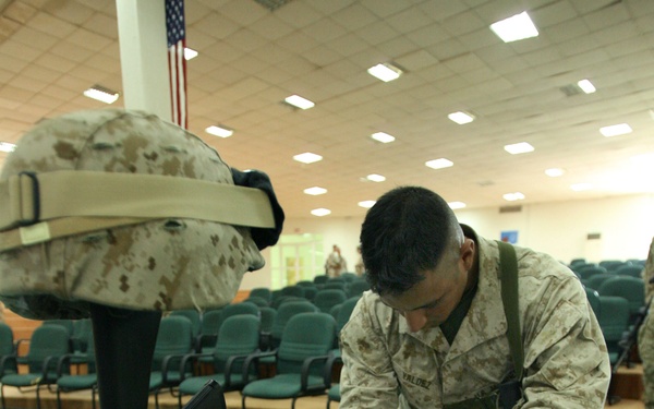 A U.S. Marine in desert MARPAT camouflage uniform kneels before a battlefield cross memorial display, holding dog tags. The memorial consists of an inverted rifle with helmet on top and combat boots at the base, a traditional military tribute to a fallen service member. The setting appears to be an indoor auditorium or chapel with rows of empty chairs and an American flag visible in the background.