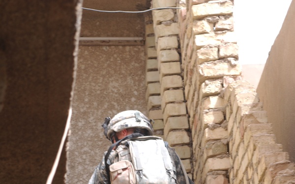 Two soldiers in US Army ACU (Army Combat Uniform) digital camouflage are navigating a narrow alleyway or stairwell between brick and concrete walls. They are fully equipped with tactical gear, body armor, helmets, and backpacks. The second soldier is carrying what appears to be a breaching tool with a red handle.