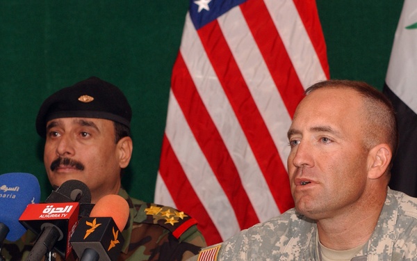 A joint press conference featuring a U.S. Army soldier identified as 'Shields' and an Iraqi military officer seated at a table with multiple media microphones. The American and Iraqi flags are visible in the background, indicating an official bilateral military briefing. Multiple Arabic-language media outlets are represented by their branded microphones in the foreground.
