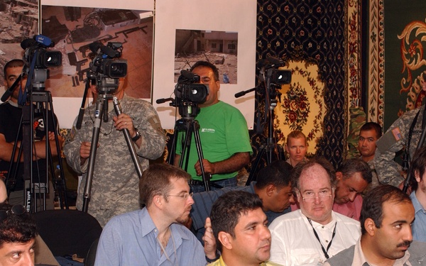 A press conference or briefing is taking place in a room decorated with ornate carpets and aerial photography displayed on the walls. Multiple journalists and cameramen are present, with several professional video cameras on tripods positioned at the back of the room. The audience consists of a mixed group of civilians and military personnel seated in rows.