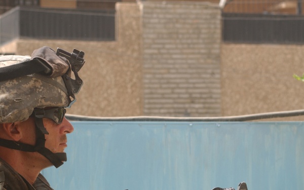 A U.S. Army soldier in full combat gear is positioned behind cover, holding an M4 carbine rifle with an optic sight attached. The soldier is wearing ACU (Army Combat Uniform) pattern camouflage, a Kevlar helmet with NVG mount, and has a U.S. flag patch and 10th Mountain Division patch visible on the left sleeve. The background shows a Middle Eastern urban environment with tan/beige concrete buildings and a blue metal gate.