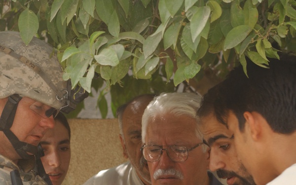 A U.S. Army soldier in full combat uniform and helmet is reviewing a document with a group of four civilian men beneath a tree. The soldier is pointing at or holding a small piece of paper while the civilians look on attentively. The setting appears to be an outdoor location, likely in a Middle Eastern country based on contextual visual cues.