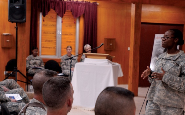 A military briefing or training session is taking place in what appears to be a chapel or meeting hall. A female soldier in ACU (Army Combat Uniform) is standing and presenting to a seated audience of fellow soldiers. A podium with a microphone and a presentation board are visible at the front of the room.