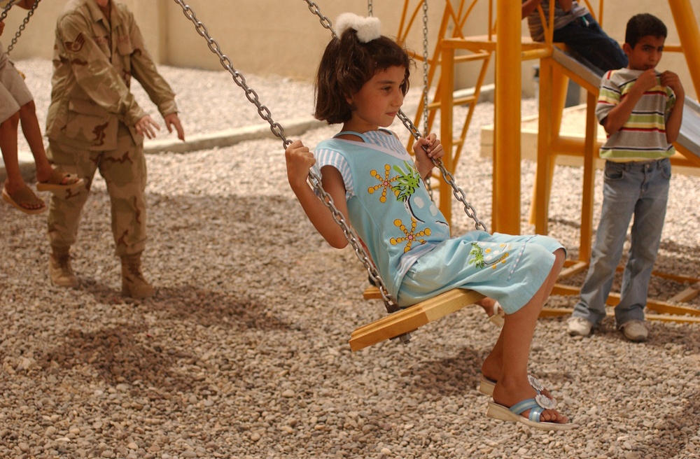 A young girl in a light blue outfit is seated on a wooden swing with metal chains in an outdoor playground area with a gravel surface. A military personnel in desert camouflage uniform is visible in the background on the left, along with other children near yellow playground equipment. The setting appears to be a newly constructed or recently installed playground, possibly in a Middle Eastern location based on contextual visual cues.