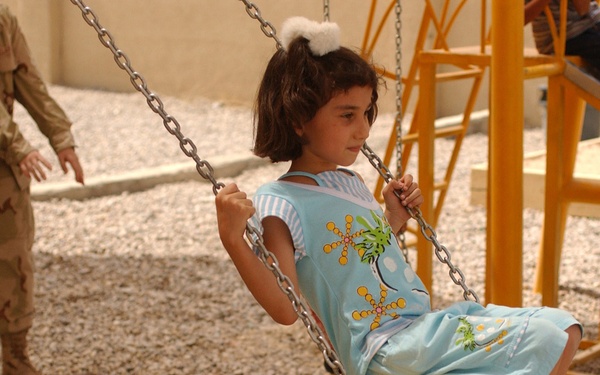 A young girl in a light blue outfit is seated on a wooden swing with metal chains in an outdoor playground area with a gravel surface. A military personnel in desert camouflage uniform is visible in the background on the left, along with other children near yellow playground equipment. The setting appears to be a newly constructed or recently installed playground, possibly in a Middle Eastern location based on contextual visual cues.