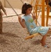 A young girl in a light blue outfit is seated on a wooden swing with metal chains in an outdoor playground area with a gravel surface. A military personnel in desert camouflage uniform is visible in the background on the left, along with other children near yellow playground equipment. The setting appears to be a newly constructed or recently installed playground, possibly in a Middle Eastern location based on contextual visual cues.