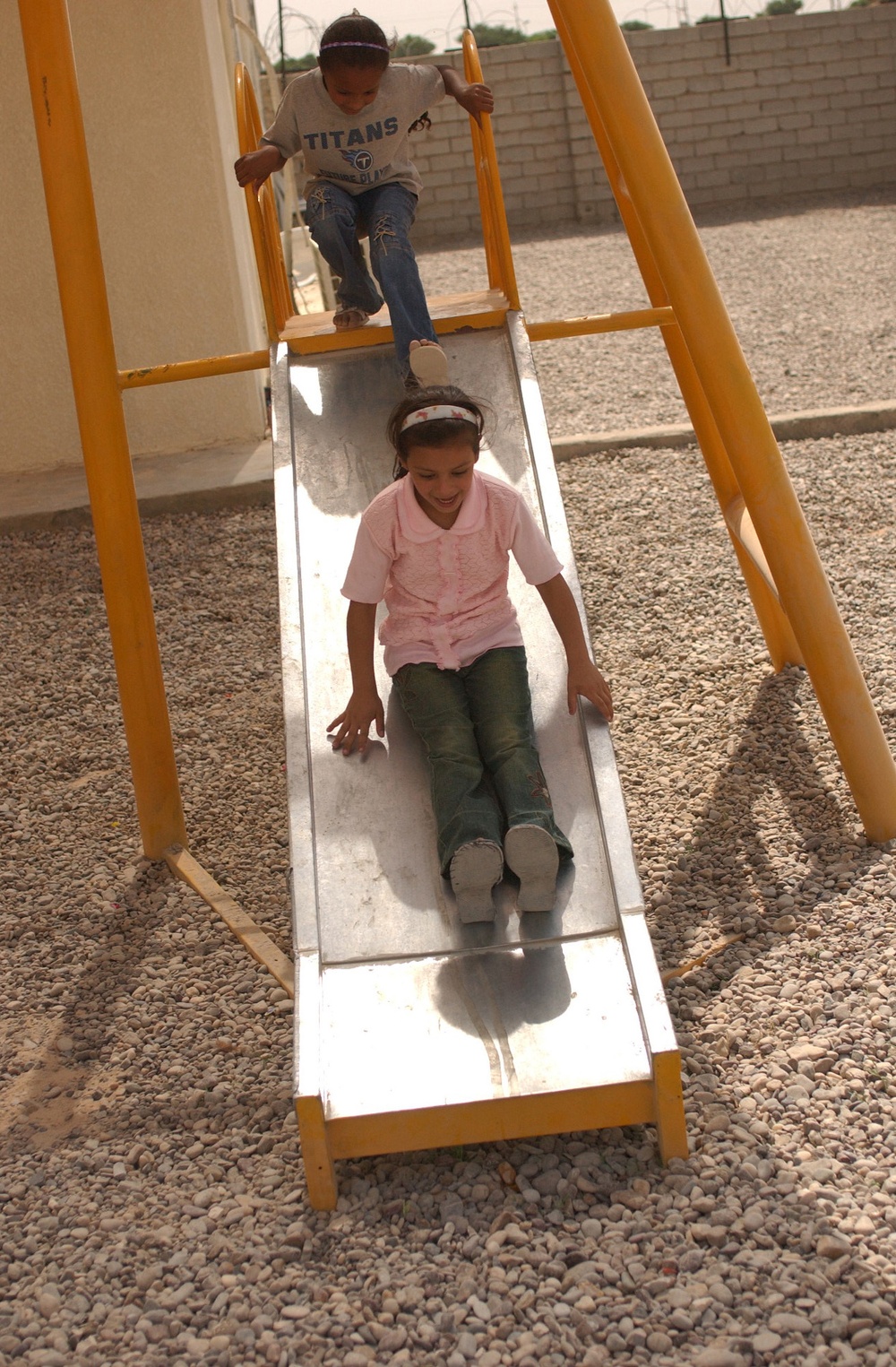 Two children are playing on a metal slide with a yellow frame in an outdoor playground area covered with gravel. A girl in a pink shirt and green pants is sliding down, while a boy wearing a 'Titans' t-shirt and jeans waits at the top. The setting appears to be a courtyard or enclosed outdoor area with a concrete block wall visible in the background.