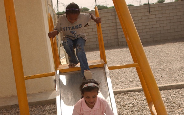 Two children are playing on a metal slide with a yellow frame in an outdoor playground area covered with gravel. A girl in a pink shirt and green pants is sliding down, while a boy wearing a 'Titans' t-shirt and jeans waits at the top. The setting appears to be a courtyard or enclosed outdoor area with a concrete block wall visible in the background.