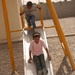 Two children are playing on a metal slide with a yellow frame in an outdoor playground area covered with gravel. A girl in a pink shirt and green pants is sliding down, while a boy wearing a 'Titans' t-shirt and jeans waits at the top. The setting appears to be a courtyard or enclosed outdoor area with a concrete block wall visible in the background.