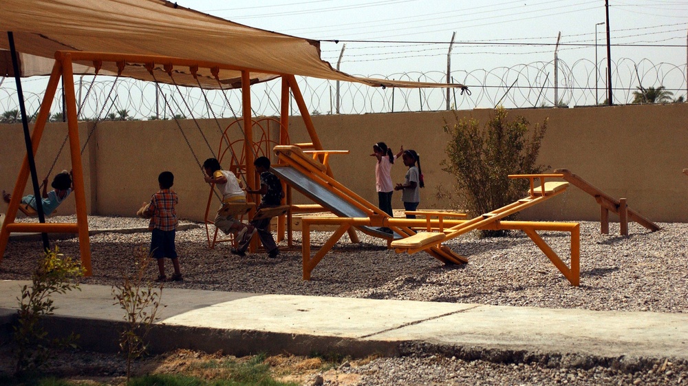 A group of children are playing on bright orange playground equipment in an outdoor area with a gravel surface. The playground is enclosed by a concrete wall topped with razor/concertina wire, suggesting a secured compound or facility. A tan shade canopy is stretched over the swing set area to provide sun protection.