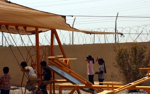 A group of children are playing on bright orange playground equipment in an outdoor area with a gravel surface. The playground is enclosed by a concrete wall topped with razor/concertina wire, suggesting a secured compound or facility. A tan shade canopy is stretched over the swing set area to provide sun protection.
