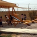 A group of children are playing on bright orange playground equipment in an outdoor area with a gravel surface. The playground is enclosed by a concrete wall topped with razor/concertina wire, suggesting a secured compound or facility. A tan shade canopy is stretched over the swing set area to provide sun protection.