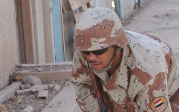 A soldier wearing desert camouflage uniform with an Iraqi flag patch is leaning over a low concrete wall or ledge, appearing to inspect or search the debris-filled area below. The soldier is equipped with a rifle with a wooden stock and is wearing sunglasses and a matching camouflage helmet. The setting appears to be an urban environment with rubble and debris visible.