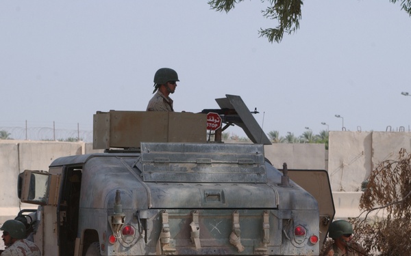 The image shows three soldiers in desert camouflage uniforms taking defensive positions around a military Humvee on a paved road. Two soldiers are crouched on either side of the vehicle at ground level with rifles raised, while a third soldier mans a position in the vehicle's turret. The setting appears to be a Middle Eastern urban or semi-urban environment, indicated by concrete blast walls, Arabic-language signage, and arid vegetation.
