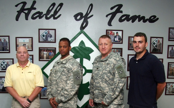 Four individuals pose for a group photo in front of a military 'Hall of Fame' display wall. Two are wearing U.S. Army ACU (Army Combat Uniform) camouflage uniforms, while two are in civilian clothing. The setting appears to be an official military facility, likely associated with the 4th Infantry Division based on the visible insignia.