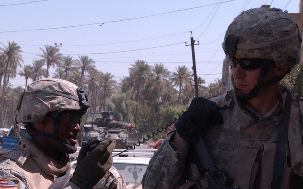 Two U.S. Army soldiers in full combat gear are engaged in communication on a street, with one soldier using a radio handset. They are wearing ACU (Army Combat Uniform) pattern camouflage, body armor, and helmets. The background shows a Middle Eastern urban environment with palm trees, utility poles, civilian vehicles, and military vehicles visible in the distance.