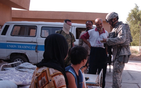 A U.S. Army soldier in ACU (Army Combat Uniform) with helmet and backpack appears to be taking notes or conducting an interview with a group of civilians outdoors. The civilians include a woman in a hijab and patterned dress seated with children, and several men standing nearby. A white van marked 'Union Trading Co.' is visible in the background along with a tan building.