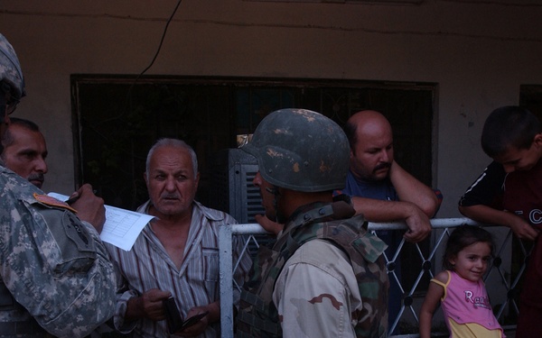 The image shows a military interaction between what appears to be a US soldier in ACU (Army Combat Uniform) and a soldier in desert camouflage alongside several civilians near a building entrance. A document or paper is being reviewed during what appears to be a checkpoint or documentation check. Civilians including a young girl and a teenage boy are present, observing the interaction.