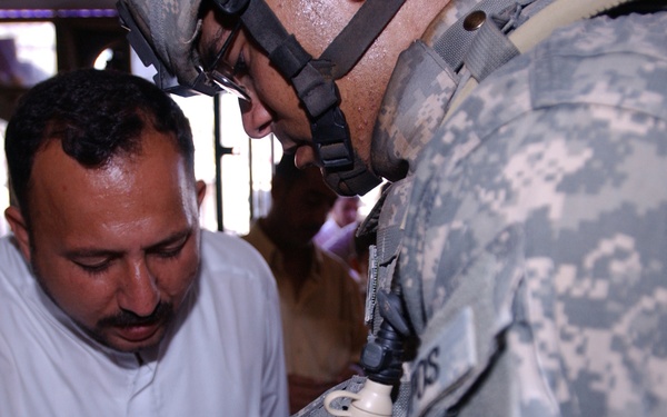 A US Army soldier in full combat gear, including ACU (Army Combat Uniform) and helmet, is reviewing a document with a civilian man wearing a white traditional garment. The interaction appears to be an administrative or identification check, with both individuals focused on paperwork. The setting appears to be an indoor or semi-covered location in what appears to be a Middle Eastern environment.