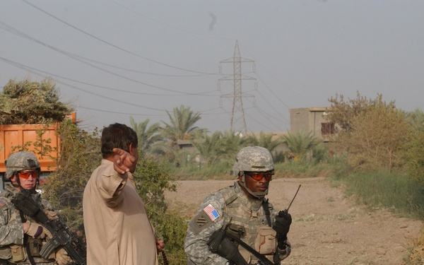 Two U.S. Army soldiers in full combat gear are interacting with a local civilian in what appears to be a rural or semi-urban area in the Middle East. The soldiers are wearing ACU (Army Combat Uniform) pattern uniforms with visible American flag patches and are armed with M4-style rifles. The scene appears to take place during a patrol or civil engagement operation.