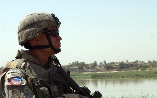 A U.S. Army soldier in full combat gear is kneeling on one knee near a concrete barrier, holding an assault rifle and scanning the surrounding area. The soldier is wearing Army Combat Uniform (ACU) digital camouflage, a Kevlar helmet with night-vision mount, body armor, and tactical gloves. A river with vegetation and buildings is visible in the background, suggesting a Middle Eastern operational environment.