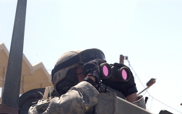 A soldier in military camouflage uniform and helmet is using binoculars with distinctive pink/magenta-coated lenses while positioned on or inside a military vehicle. The setting appears to be an urban environment with tan/beige buildings visible in the background. The image captures a surveillance or reconnaissance activity in what appears to be a Middle Eastern or arid-climate location.