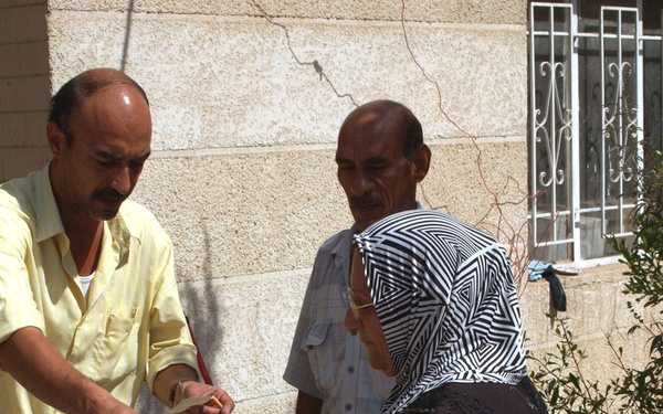 Three individuals are gathered outside a stone building in what appears to be a distribution or exchange of goods. A man in a yellow shirt is handing items to a woman dressed in a black abaya and black-and-white patterned hijab, while a second bald man in a light blue shirt observes in the background. A yellow plastic bag and a cardboard box are visible, suggesting a food or supply distribution activity.