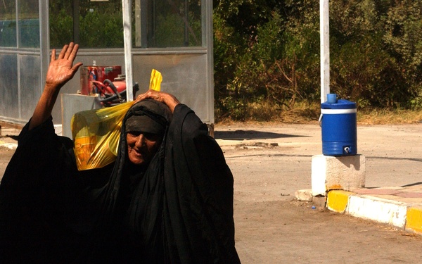 An elderly woman dressed in a black abaya and hijab is walking in an outdoor paved area, raising one hand as if waving or gesturing. She is carrying a yellow bag on her shoulder. In the background, a man in a yellow shirt and jeans walks away, and a glass shelter structure is visible along with trees and a blue water cooler.
