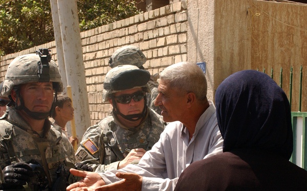 The image shows U.S. Army soldiers in full combat gear engaging in a conversation with local civilians, likely during a patrol or community interaction mission. The soldiers are wearing ACU (Army Combat Uniform) digital camouflage and helmets, while the civilians include an older man gesturing with open hands, a woman in a dark hijab, and a child. The setting appears to be a residential street with brick walls and a green metal fence.