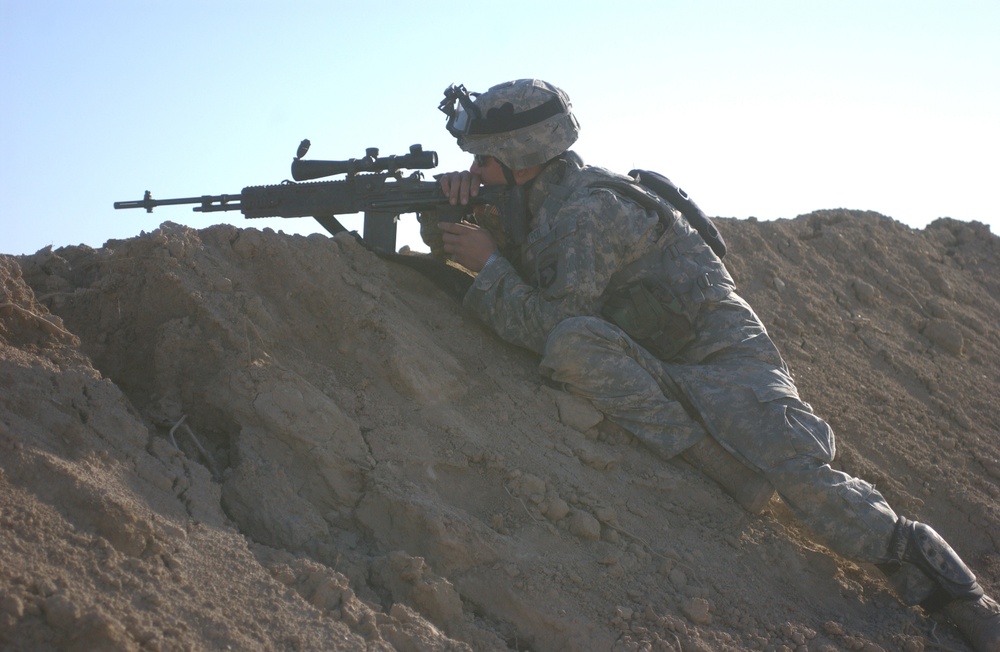 A soldier in US Army Universal Camouflage Pattern (UCP) uniform is positioned prone on a sandy berm, aiming a scoped semi-automatic rifle. The soldier is wearing a combat helmet with what appears to be a night vision device mount and tactical gear. The arid, desert-like terrain and clear sky suggest an operational or training environment in a dry region.