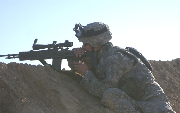 A soldier in US Army Universal Camouflage Pattern (UCP) uniform is positioned prone on a sandy berm, aiming a scoped semi-automatic rifle. The soldier is wearing a combat helmet with what appears to be a night vision device mount and tactical gear. The arid, desert-like terrain and clear sky suggest an operational or training environment in a dry region.