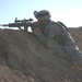 A soldier in US Army Universal Camouflage Pattern (UCP) uniform is positioned prone on a sandy berm, aiming a scoped semi-automatic rifle. The soldier is wearing a combat helmet with what appears to be a night vision device mount and tactical gear. The arid, desert-like terrain and clear sky suggest an operational or training environment in a dry region.
