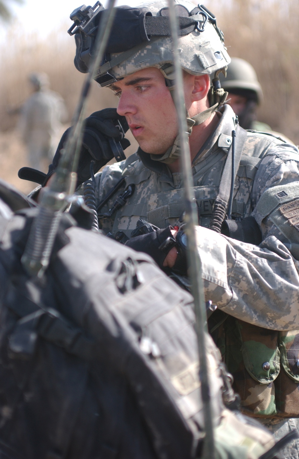 A U.S. Army soldier in ACU (Army Combat Uniform) digital camouflage is focused on equipment, appearing to operate or inspect a radio/communication device. He is wearing a combat helmet with a night vision goggle mount and tactical headset. At least two other military personnel are visible in the background.