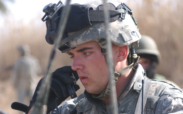 A U.S. Army soldier in ACU (Army Combat Uniform) digital camouflage is focused on equipment, appearing to operate or inspect a radio/communication device. He is wearing a combat helmet with a night vision goggle mount and tactical headset. At least two other military personnel are visible in the background.
