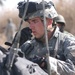 A U.S. Army soldier in ACU (Army Combat Uniform) digital camouflage is focused on equipment, appearing to operate or inspect a radio/communication device. He is wearing a combat helmet with a night vision goggle mount and tactical headset. At least two other military personnel are visible in the background.