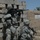 Two soldiers in U.S. Army ACU (Army Combat Uniform) digital camouflage are positioned against a concrete block wall in what appears to be an arid, Middle Eastern environment. One soldier is standing and aiming an M4 carbine with an optic sight, while a second soldier kneels beside them also holding a weapon. The setting features mud-brick and concrete block construction typical of Iraq or Afghanistan.